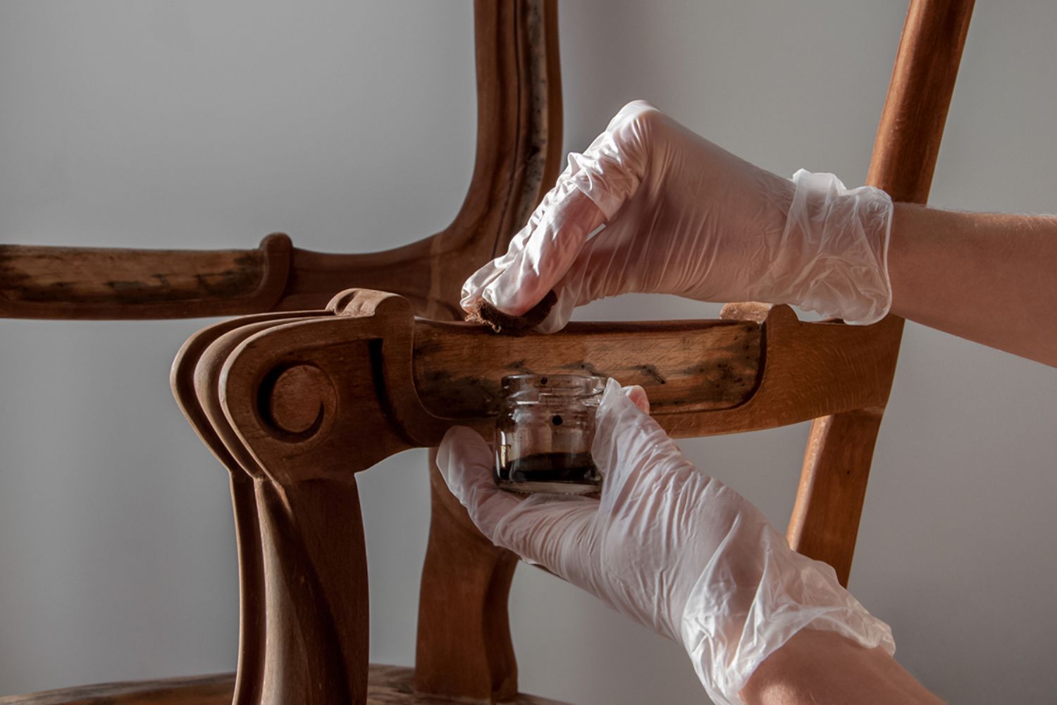 A person in white gloves applies wood finish from a jar to enhance the intricate design of a wooden chair.
