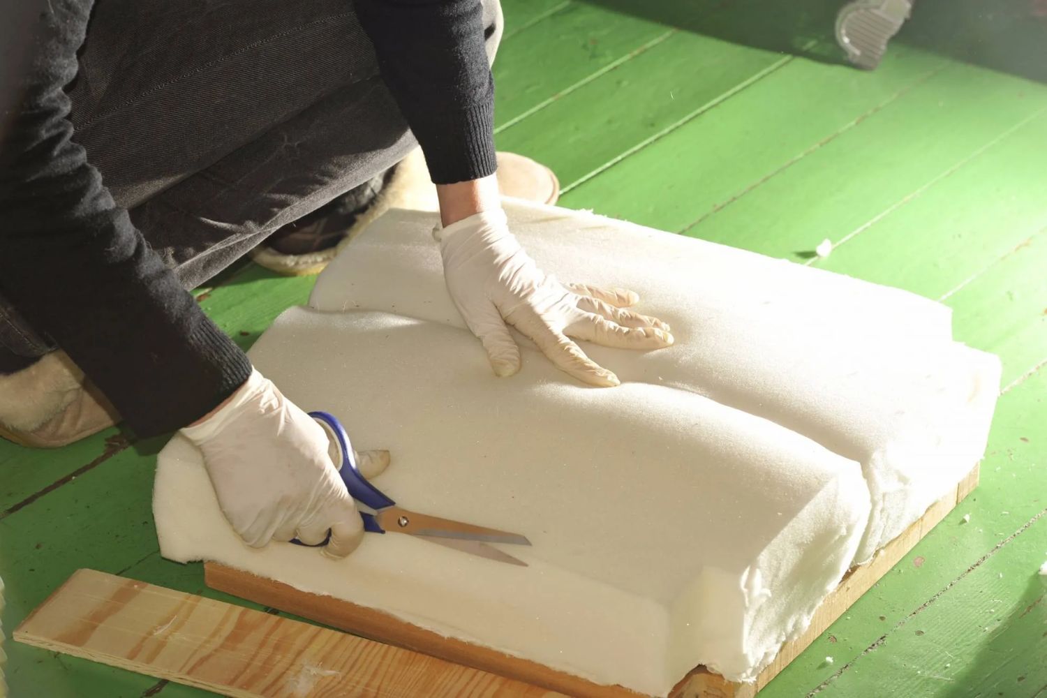 A person in gloves cuts into a large block of soft material with scissors, working on a green wooden surface.