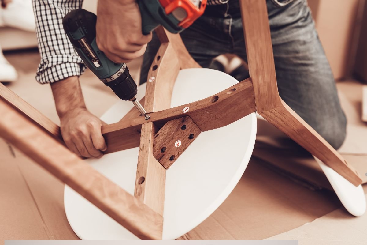 A person assembling a wooden table using a power drill, securing the legs to a round white tabletop on a cardboard surface.